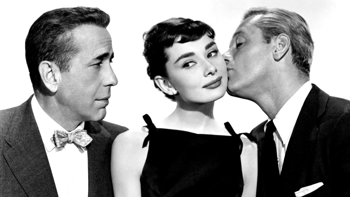 A woman stands between two men. One stares at her longingly while the other kisses her cheek.