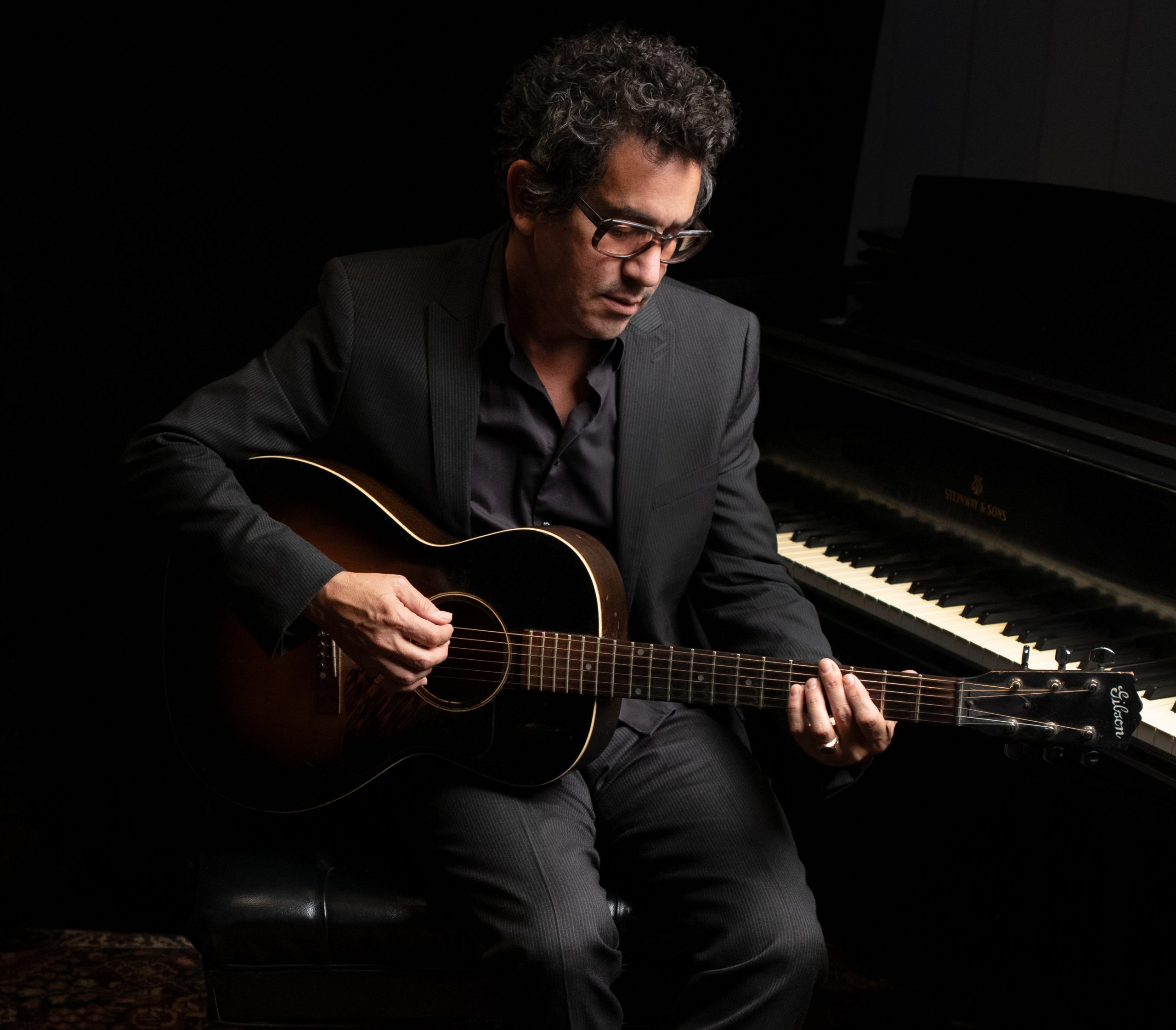 A man sitting at a piano bench with an acoustic guitar