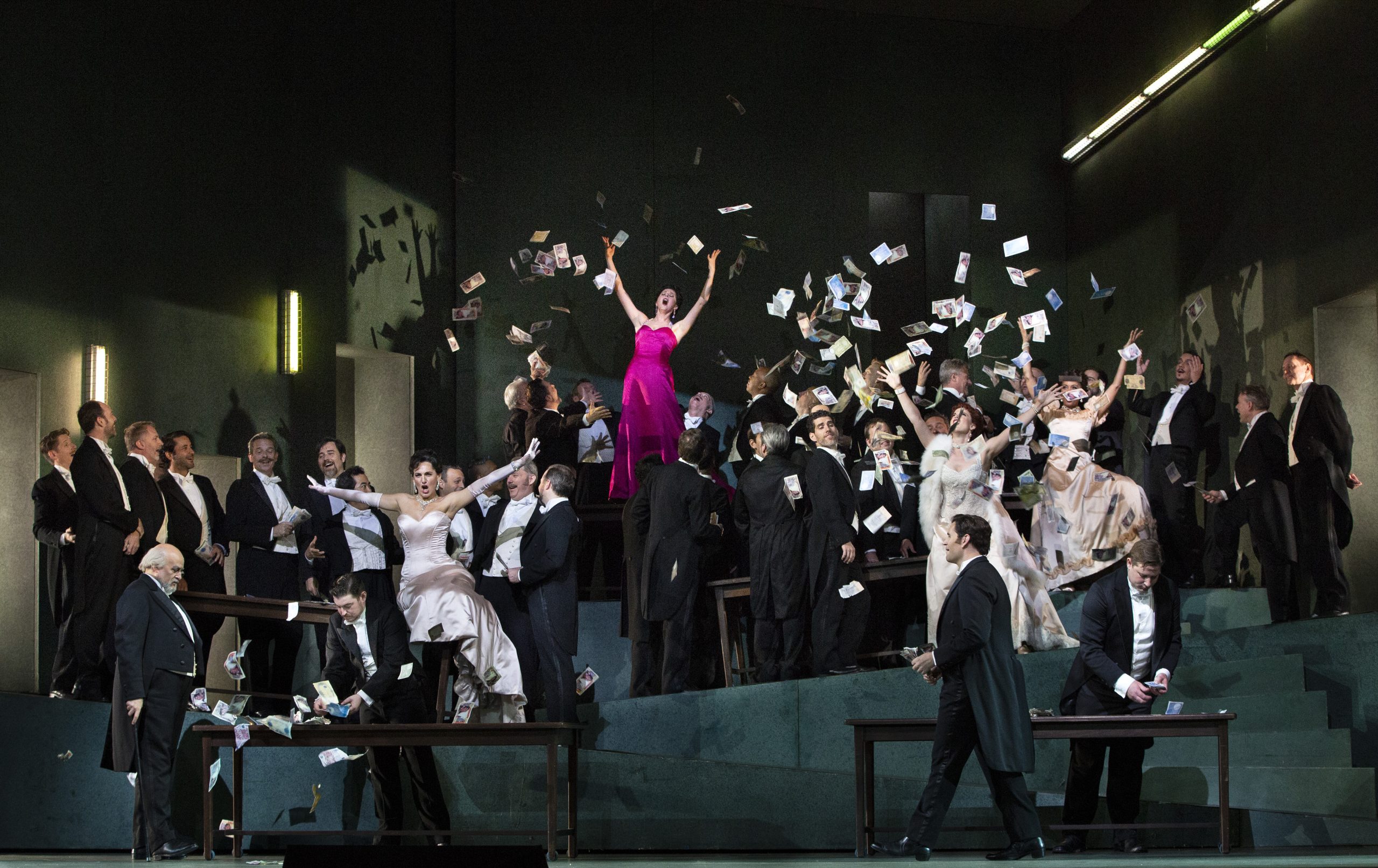 An opera staging of a group of formally dressed people, with a woman in a pink dress tossing money