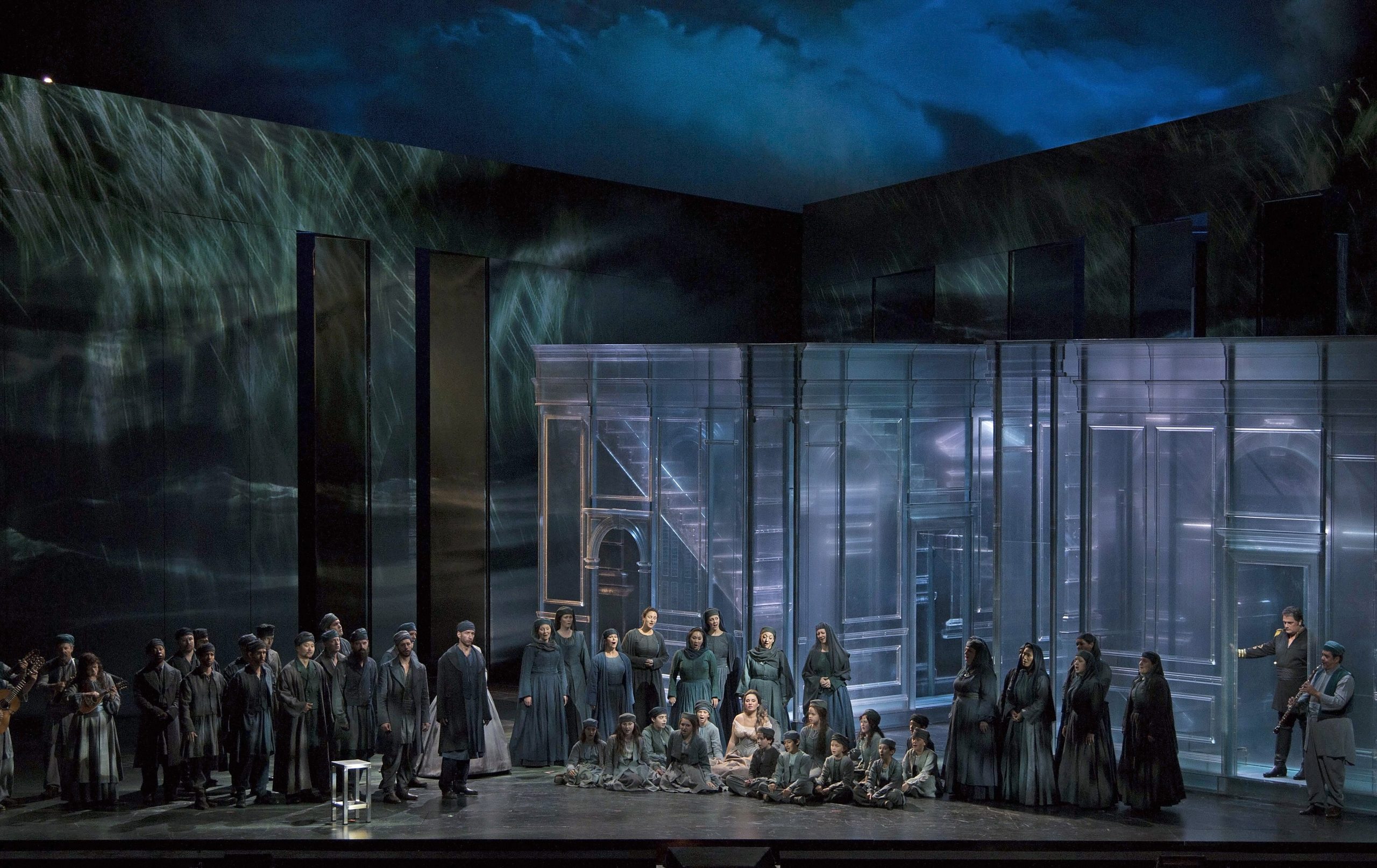An opera staging with a large group of individuals gathered around, with children seated on the ground and adults standing around them