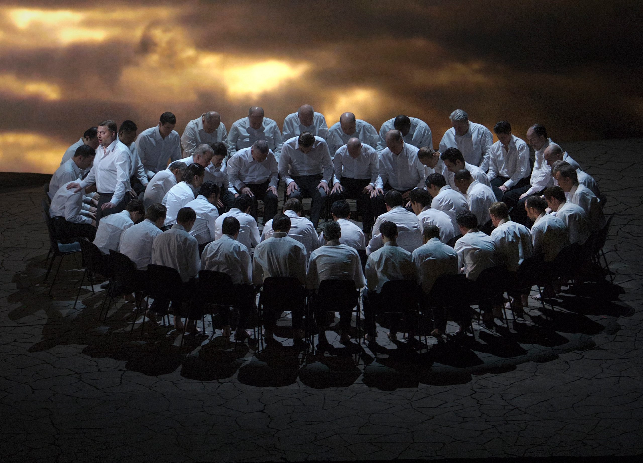 An opera staging of a group of men seated in a circle with a gloomy sky behind them